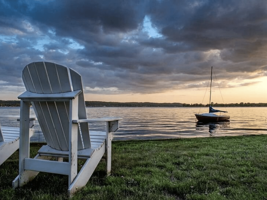 Two white Adirondack Chairs next to the lake at Canfield House in Onekema with a small sailboat moored