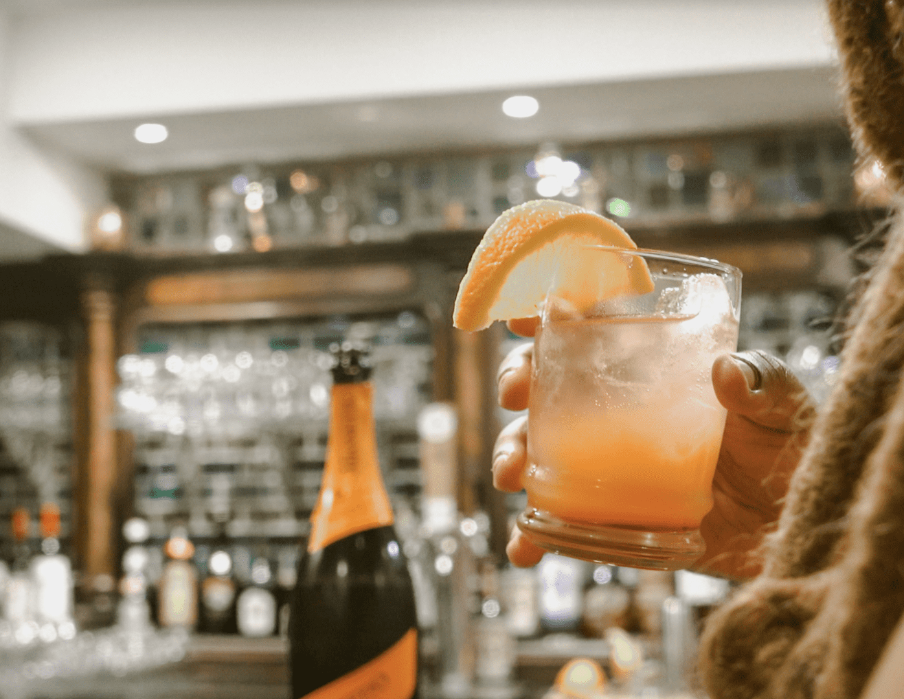 Woman holding a handcrafted cocktail with a lemon and bottle of Prosecco in the background