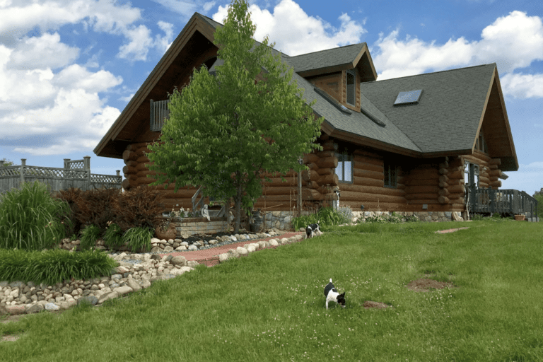 A log cabin on top of a hill with a small dog in the yard.