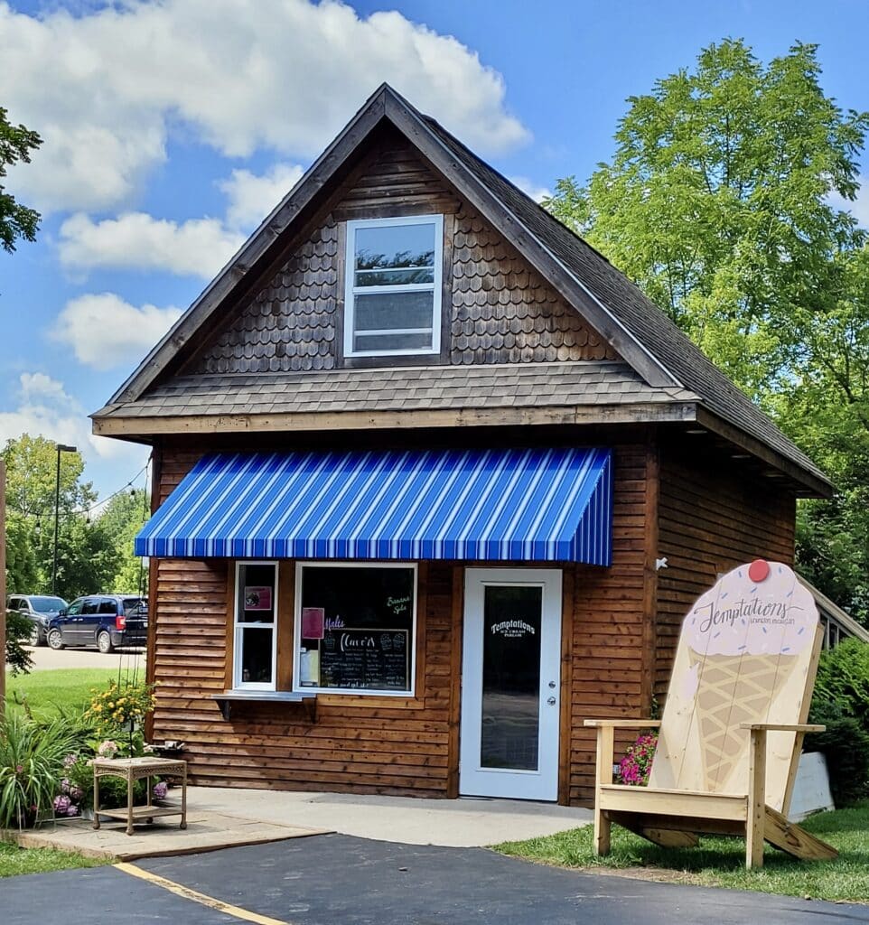 Scoops of Lexington ice Cream shop with a large yellow chair beside the quaint small brown building with a blue awning.